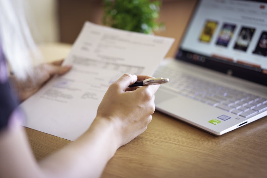 Close-up of a hand holding a pen and paper near a laptop, suggesting online shopping or document review.