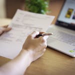 Close-up of a hand holding a pen and paper near a laptop, suggesting online shopping or document review.