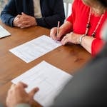Two adults sign documents on a wooden table at a business meeting, emphasizing teamwork.